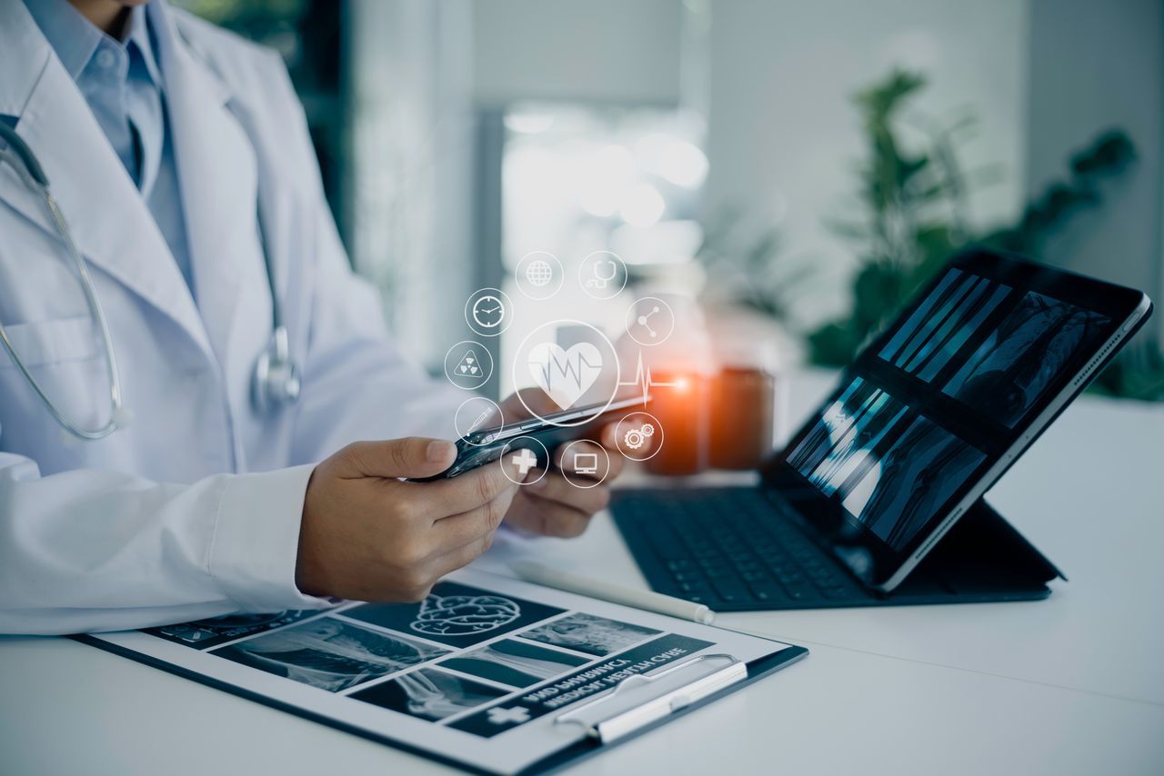 Arzt am Arbeitsplatz mit virtuellen Gesundheitsanwendungen auf dem Handy und medizinischen Diagnosebildern auf dem Laptop.