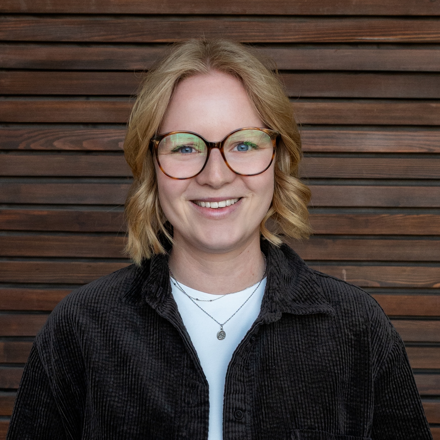 DSC_3161_1x1 Smiling woman with blonde hair and round glasses in front of a wooden wall stylized backdrop.