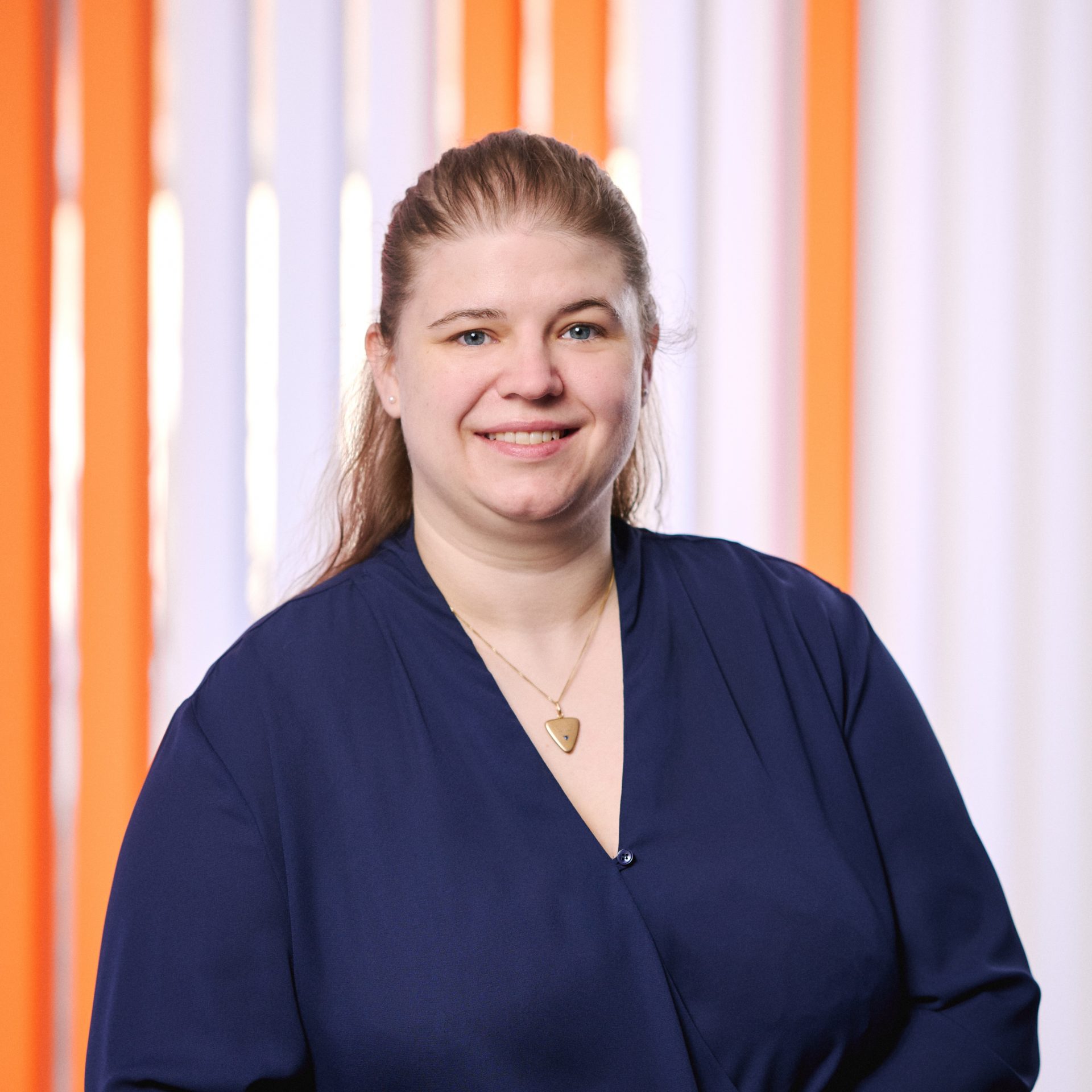 Marina Iftner 1x1 Smiling person in a navy top, with orange and white striped background behind them.