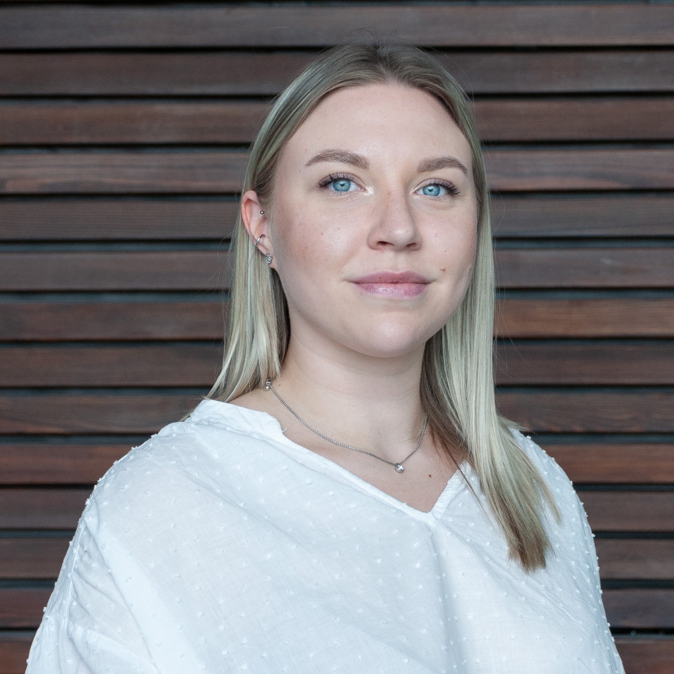 Thumbnail_Medical Valley EMN e V_Julia Huber Portrait of a smiling young woman with blonde hair and blue eyes, wearing a white textured blouse, against a wooden slat wall.